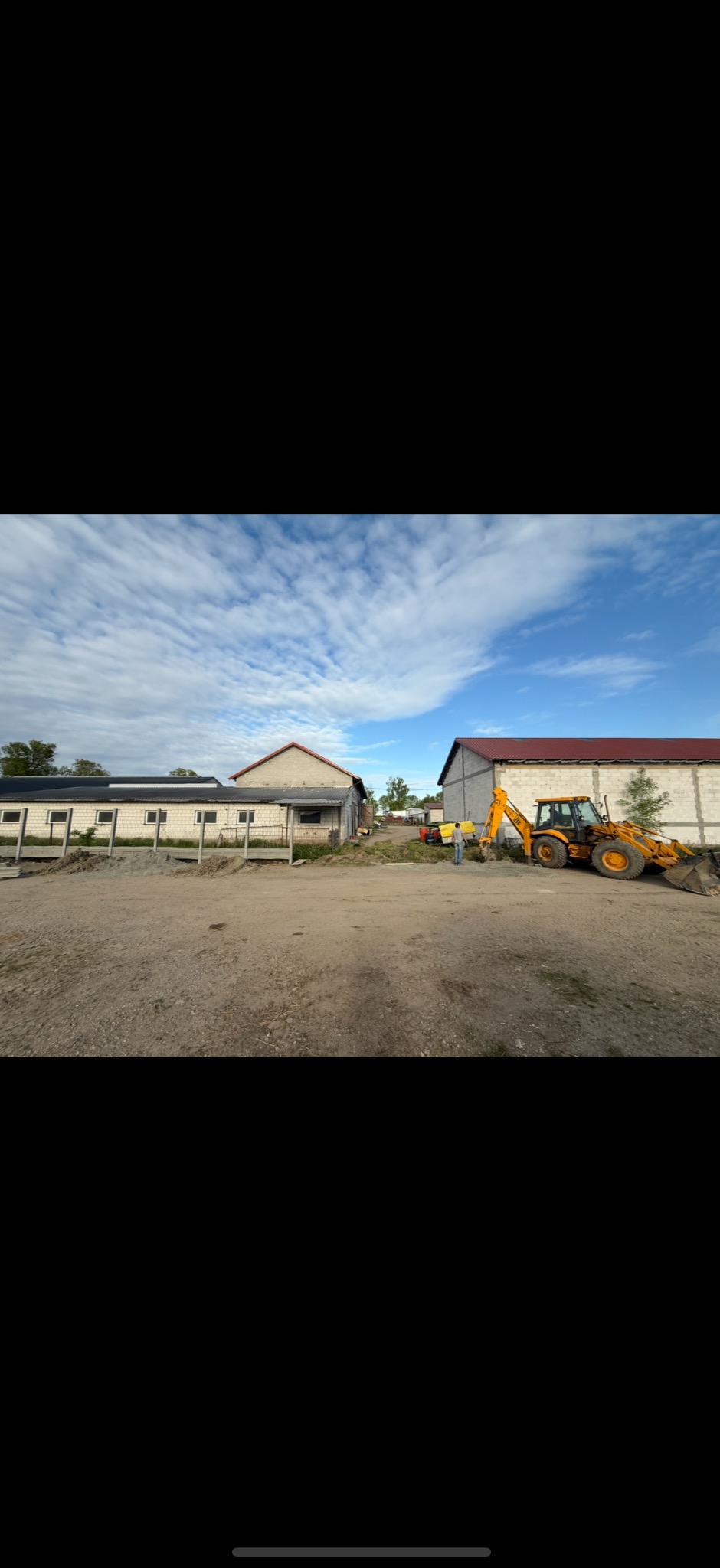 Żółta koparko-ładowarka na placu budowy z budynkami w tle i błękitnym niebem. Prace ziemne w toku, widoczny operator i teren przygotowywany pod inwestycję.