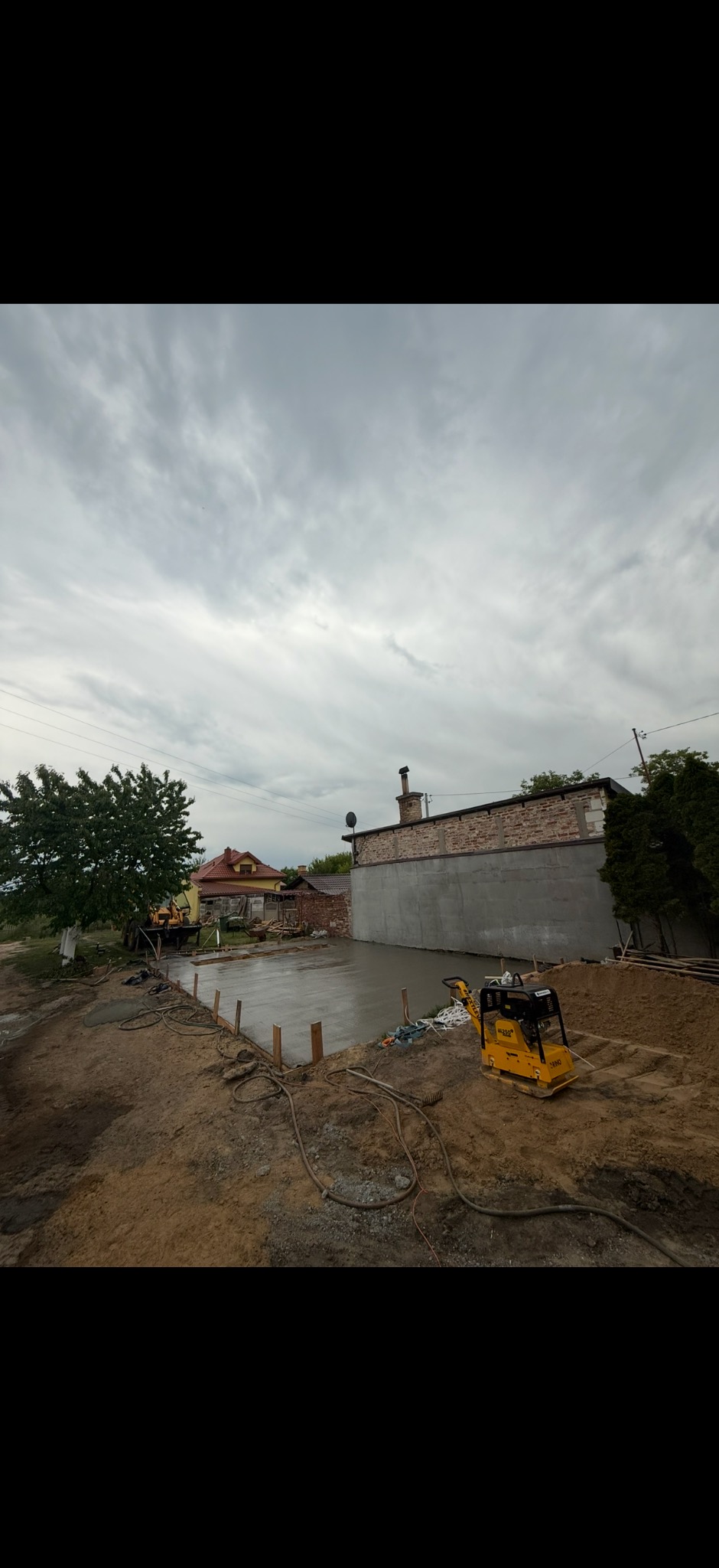 Wylany fundament pod budowę z żółtą zagęszczarką obok. Widoczny szalunek, budynek w tle i pochmurne niebo.