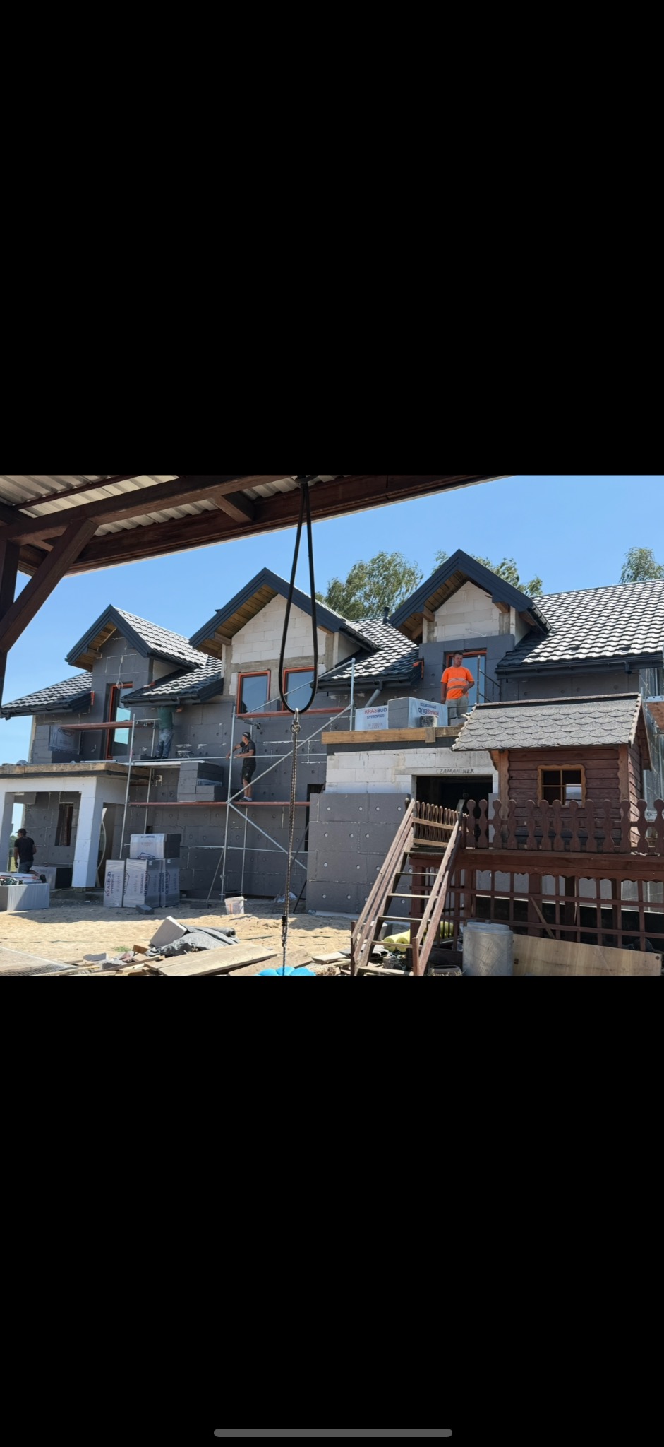 Budowa domu jednorodzinnego w trakcie ocieplania elewacji, widoczne rusztowania i pracownicy. Na pierwszym planie domek dla dzieci z drewnianym ogrodzeniem.