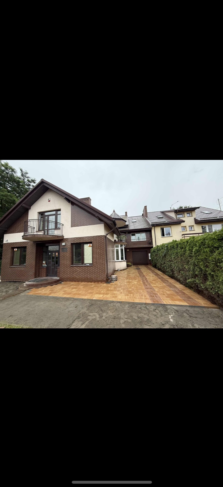 Nowa, brązowo-żółta kostka brukowa przed domem z cegły. Ułożona wzorem w pasy, prowadzi do garażu. Widoczne ślady ziemi po bokach. Dom dwukondygnacyjny z balkonem.