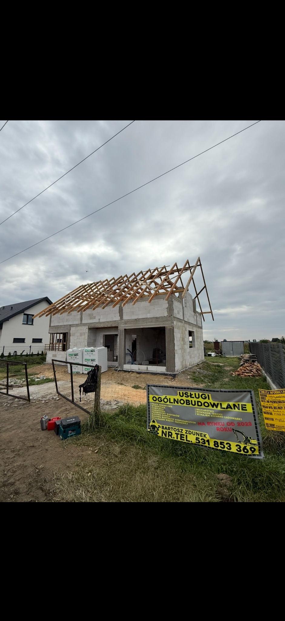 Budowa domu z pustaków, widoczne drewniane elementy konstrukcji dachu. Na pierwszym planie baner reklamowy firmy budowlanej. Pochmurne niebo w tle.