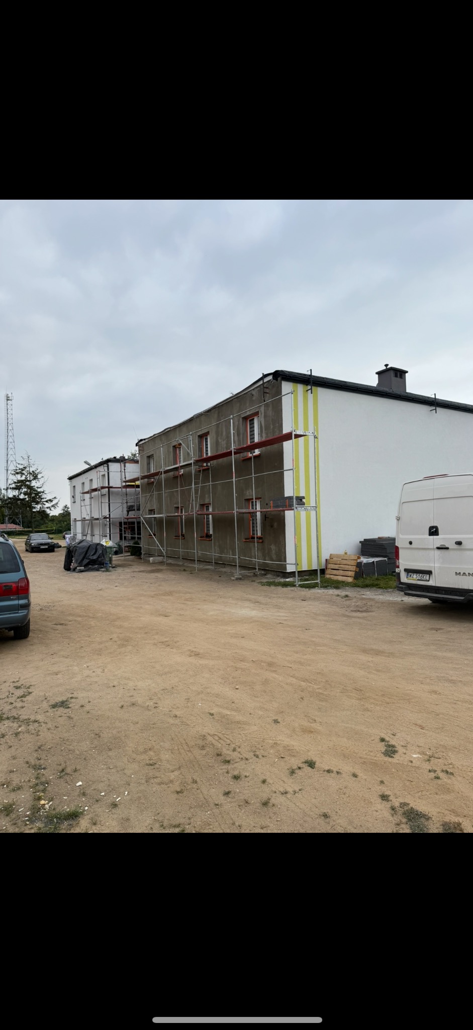 Budynek w trakcie ocieplania elewacji, widoczne rusztowania i częściowo otynkowana ściana z żółtymi pasami. Biały bus i samochody na placu budowy.
