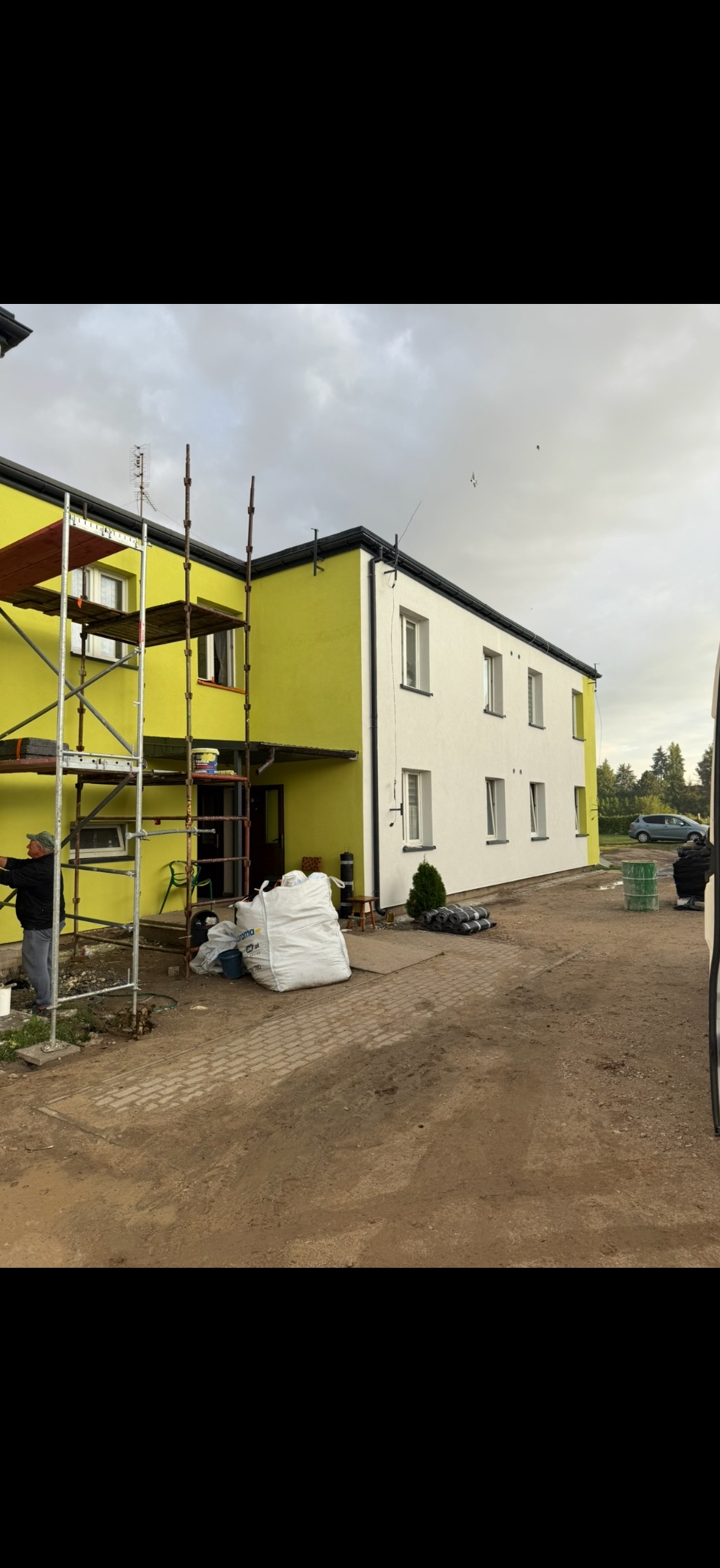 Dwukondygnacyjny budynek częściowo otynkowany na biało, część w kolorze żółtym, z rusztowaniem i workami materiałów budowlanych przed wejściem.