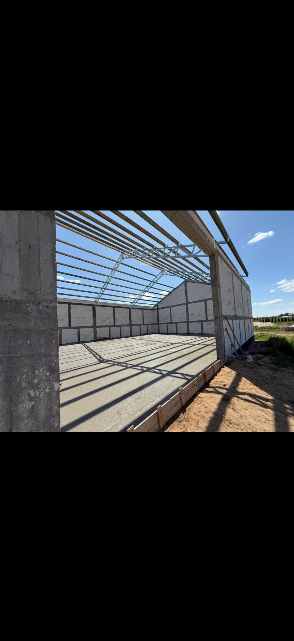 Wnętrze surowego, jasnego budynku w trakcie budowy. Widoczne ściany z betonu komórkowego, wylana posadzka i metalowa konstrukcja dachu. Zewnętrzne otoczenie z zielenią w tle.