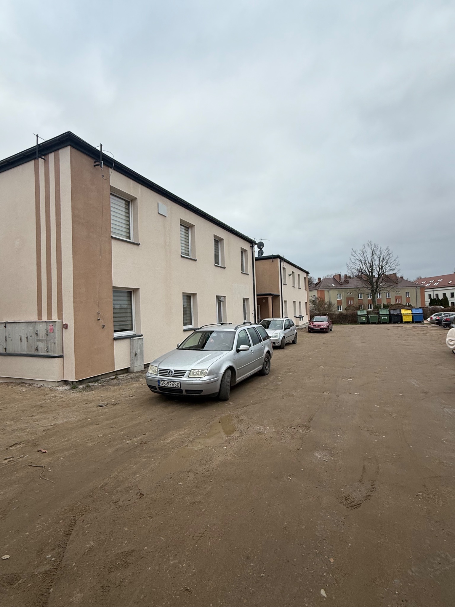 Odnowiona elewacja budynku z beżowymi pasami, widok z parkingu z samochodami w pochmurny dzień. Świeżo pomalowane ściany i uporządkowany teren.