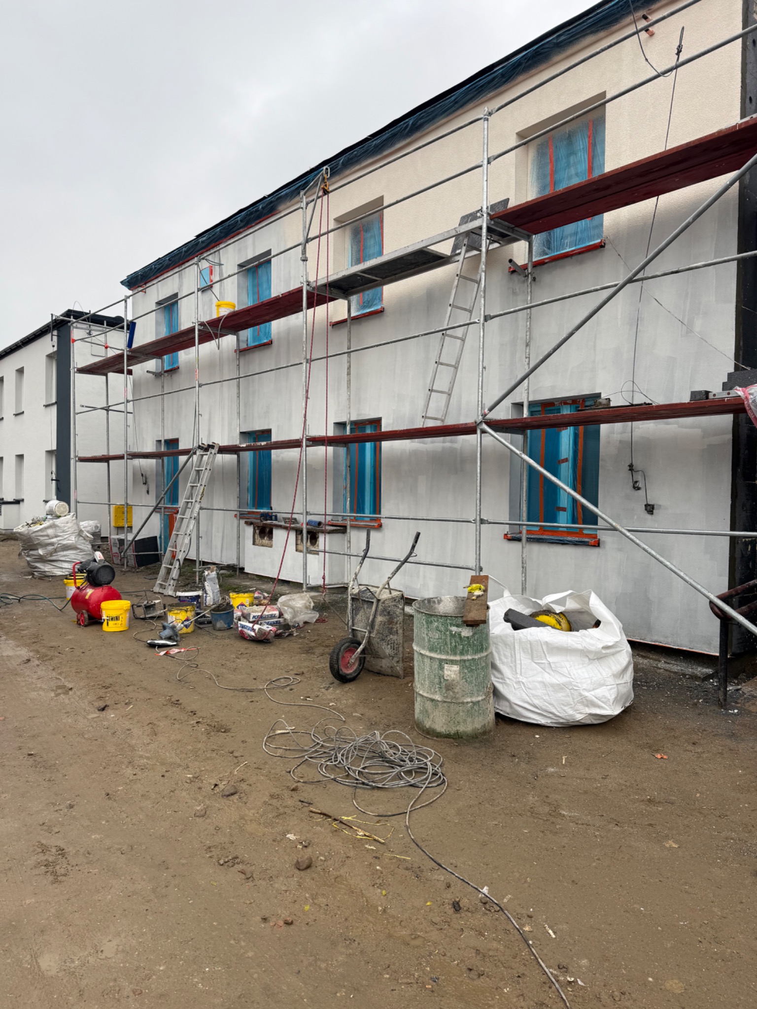 Budynek w trakcie prac elewacyjnych: rusztowania, okna zabezpieczone folią, widoczne narzędzia i materiały budowlane. Świeżo nałożony tynk w jasnym odcieniu.