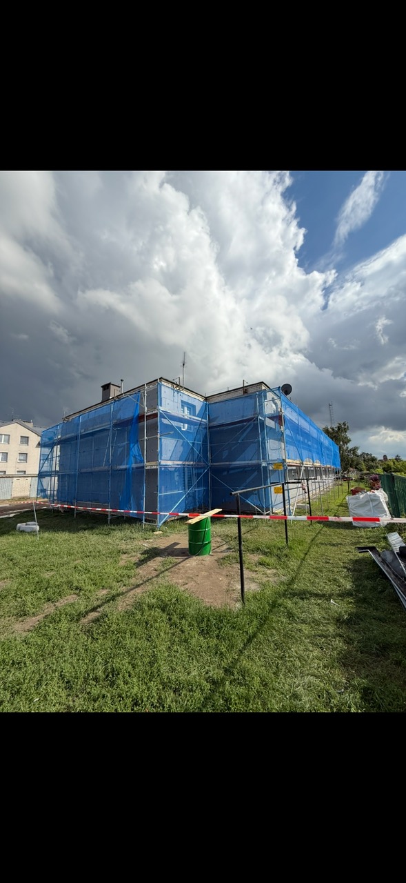 Budynek w rusztowaniach, zabezpieczony niebieską siatką, przygotowany do prac elewacyjnych. Widoczne ogrodzenie i elementy zabezpieczające teren budowy.