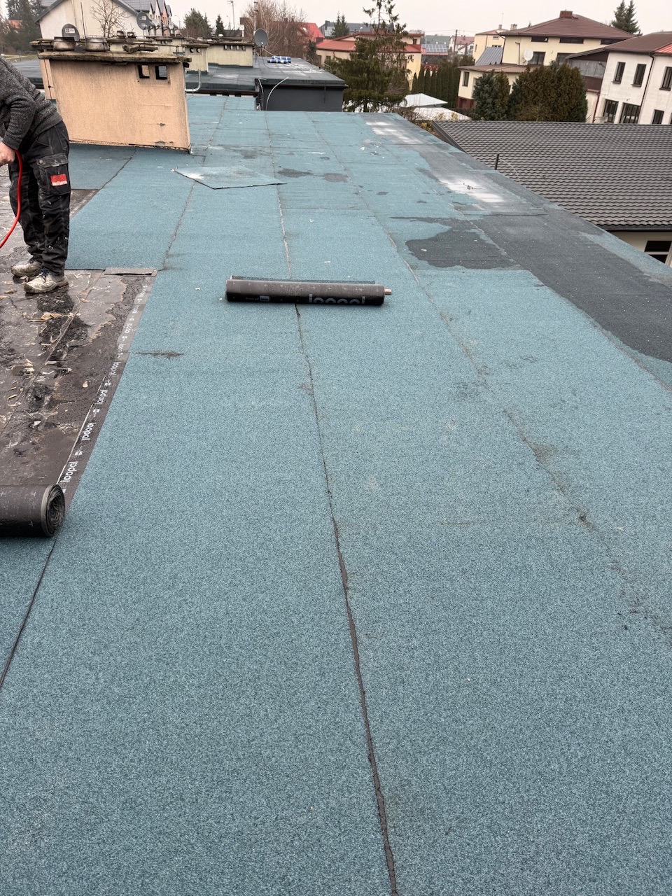 Prace na dachu płaskim: fragment pokrycia z papy termozgrzewalnej w kolorze niebiesko-szarym, częściowo ułożonej, z widocznym pracownikiem i rolką materiału.