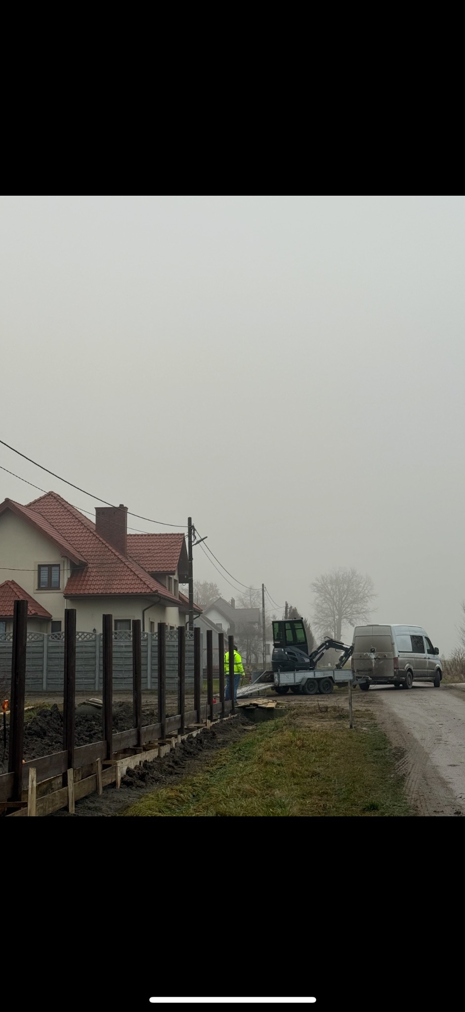 Montaż ogrodzenia panelowego z drewnianymi słupkami i podmurówką. W tle dom jednorodzinny i mini koparka na przyczepie. Prace ziemne w toku, pochmurny dzień.