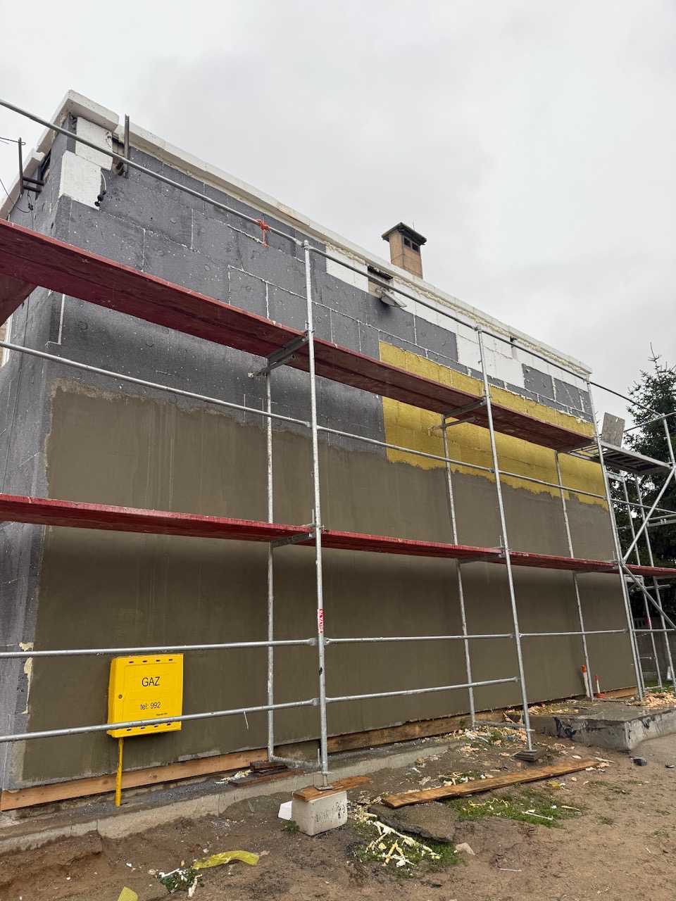 Ściana budynku w trakcie prac elewacyjnych: ocieplenie styropianem, fragment tynku, rusztowanie. Żółta skrzynka gazowa na ścianie. Widok z dołu, pochmurne niebo.
