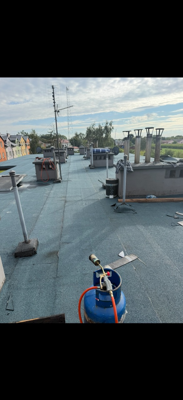 Niebieski dach pokryty papą termozgrzewalną z kominami i anteną. W tle kolorowe budynki. Palnik dekarski na pierwszym planie.