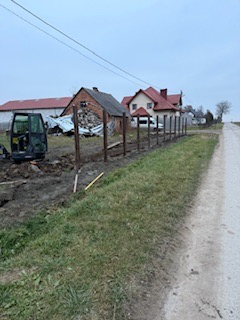 Montaż ogrodzenia z drewnianych słupków w wiejskim krajobrazie. Widoczny mini-koparka i przygotowany teren pod ogrodzenie. Pochmurne niebo.