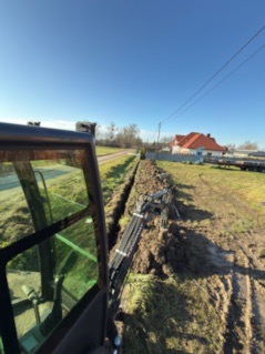 Wykopany wąski rów w ziemi, widok z kabiny koparki na tle błękitnego nieba i zabudowań w oddali. Prace ziemne w słoneczny dzień.