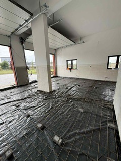 Zbrojenie podłoża wewnątrz budynku z bramą garażową, widoczne okna i kolumna nośna. Siatka zbrojeniowa na czarnej folii, przygotowanie do wylewki.