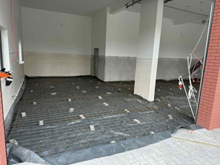 Zbrojenie pod wylewkę w garażu, siatka zbrojeniowa na czarnej folii, betonowe podkładki dystansowe, widoczne przewody elektryczne i brama garażowa.