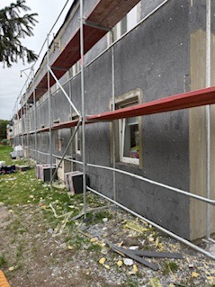 Elewacja budynku w trakcie prac: szare ocieplenie, rusztowania, okno z widoczną firanką. Fragmenty styropianu na trawie. Ujęcie z dołu, lekko zniekształcona perspektywa.