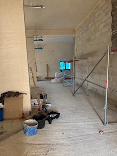 Wnętrze w trakcie prac wykończeniowych: surowy stan, widoczne rusztowania, narzędzia malarskie i przygotowane ściany. Okno w głębi kadru.