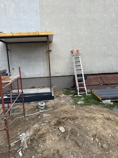 Budowa ganku przy ścianie budynku z drabina i rusztowaniem. Widoczny fragment ocieplenia. Prace w toku, ziemia wokół. Ujęcie z dołu, lekko z prawej strony.