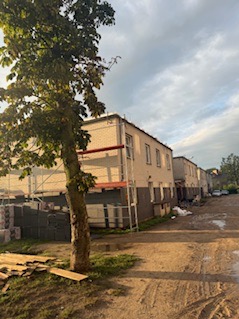 Budynek w trakcie prac elewacyjnych z rusztowaniem, widok z dołu, z boku drzewo, w tle pochmurne niebo. Częściowo ocieplona ściana, widoczne materiały budowlane.