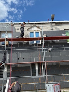 Praca na rusztowaniu: ocieplanie elewacji budynku styropianem grafitowym. Widoczne okna, fragment dachu i niebo. Jeden pracownik na górze, drugi na dole.