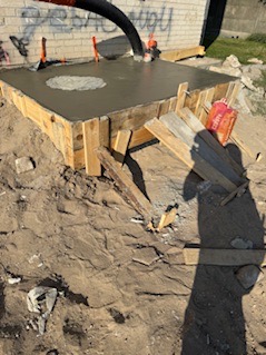 Zalewanie betonem szalunku fundamentu pod budowę. Widoczny wąż pompy do betonu i drewniany szalunek. Tło stanowi mur z graffiti.