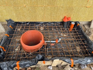 Zbrojenie fundamentu pod budynek, widoczne pręty stalowe, folia ochronna i rury kanalizacyjne. Ocieplenie ściany budynku w tle. Ujęcie z góry.