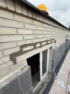 Fragment ściany z jasnej cegły z oknem, częściowo ocieplonej szarym styropianem. Na górze żółty kask budowlany. Widoczne ślady prac murarskich.