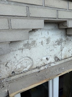 Uszkodzona elewacja z cegły, widoczne ubytki i pęknięcia. Fragment okna w dolnej części kadru. Nierówna powierzchnia, brak spójności wizualnej.