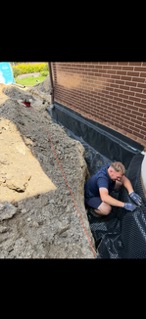 Pracownik w wykopie układa folię kubełkową przy fundamencie ceglanego budynku. Widoczne zabezpieczenie przeciwwilgociowe i drenaż. Prace ziemne w toku.