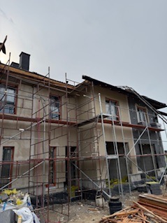 Dwukondygnacyjny dom w trakcie prac elewacyjnych, z widocznymi rusztowaniami i fragmentami ocieplenia. Część fasady pokryta szarym tynkiem, reszta przygotowana do dalszych prac.