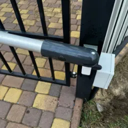 Siłownik automatyczny do bramy skrzydłowej, zamontowany na czarnym, metalowym słupku i bramie, widoczne kostki brukowe w tle.