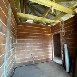 Surowy stan wnętrza: mury z pustaków ceramicznych, drewniana konstrukcja dachu, instalacja elektryczna w peszlach przy podłodze. Widoczne materiały budowlane.