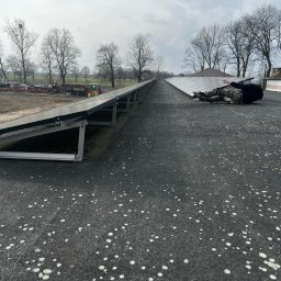 ElektroTechInstal Patryk Kowalczyk - Instalacja paneli słonecznych na dachu budynku gospodarczego. Osoba w kombinezonie montuje panele. W tle widoczne drzewa i sprzęt rolniczy.