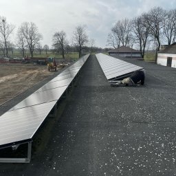 ElektroTechInstal Patryk Kowalczyk - Instalacja paneli słonecznych na dachu budynku gospodarczego. Pracownik w trakcie montażu, widoczne rzędy paneli i sprzęt rolniczy w tle. Pochmurny dzień.