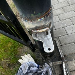 ElektroTechInstal Patryk Kowalczyk - Spawanie uchwytu do słupka bramy, widoczne ślady spawania i tęczowe przebarwienia. Obok leżą robocze rękawice i narzędzia. Tło z kostki brukowej i trawy.