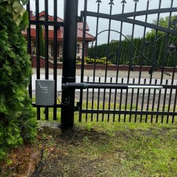 ElektroTechInstal Patryk Kowalczyk - Czarna, metalowa brama z zamontowanym siłownikiem automatyki, widoczny sterownik FORCE, na tle zieleni i domu z czerwoną dachówką w pochmurny dzień.