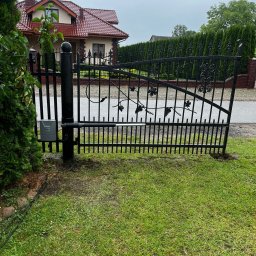 ElektroTechInstal Patryk Kowalczyk - Czarna, metalowa brama z automatyką na tle domu z czerwoną dachówką i zieleni. Widoczny siłownik bramy. Pochmurne niebo.