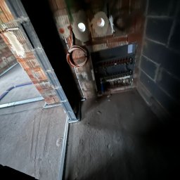 ElektroTechInstal Patryk Kowalczyk - Wnętrze w trakcie prac instalacyjnych: widoczne przewody elektryczne, rozdzielnica i elementy konstrukcyjne sufitu podwieszanego. Surowy stan, cegła i beton.