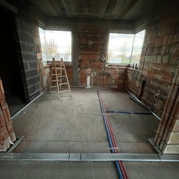 ElektroTechInstal Patryk Kowalczyk - Surowy stan wnętrza z instalacjami podtynkowymi i rurami w podłodze. Widoczne puszki elektryczne w ścianach z cegły i drabina. W tle okna.