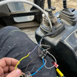 Elektryk łączy przewody wewnątrz kabiny maszyny budowlanej. Widoczne złącza, przewody w różnych kolorach i narzędzia. Praca w trudnych warunkach.