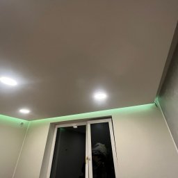 ElektroTechInstal Patryk Kowalczyk - Nowoczesne oświetlenie sufitowe z zielonymi paskami LED nad oknem, uzupełnione punktowymi lampami. Minimalistyczny design wnętrza z jasnymi ścianami.