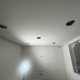 ElektroTechInstal Patryk Kowalczyk - Białe wnętrze z przygotowanymi otworami na oświetlenie sufitowe. Widoczna drabina i lampa budowlana. Minimalistyczny design, surowy stan wykończenia.