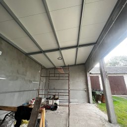 ElektroTechInstal Patryk Kowalczyk - Montaż sufitu podwieszanego z metalową konstrukcją w garażu. Widoczny rusztowanie, narzędzia i lampa. Surowy beton na ścianach. Ujęcie z dołu, szeroki kadr.