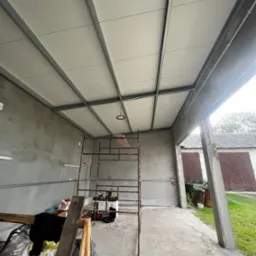 Montaż sufitu podwieszanego z metalową konstrukcją w garażu. Widoczny rusztowanie, narzędzia i lampa. Surowy beton na ścianach. Ujęcie z dołu, szeroki kadr.