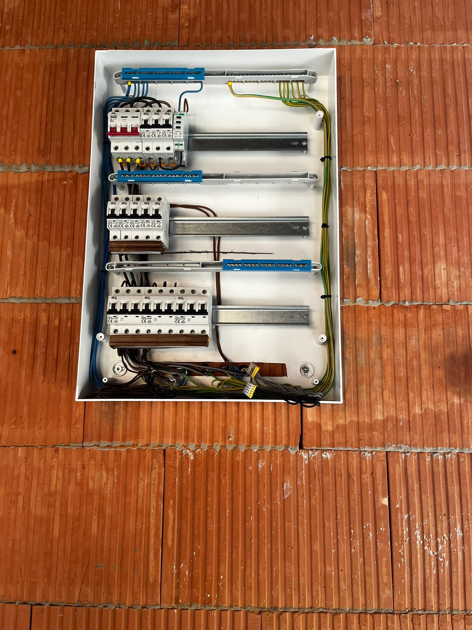 Rozdzielnia elektryczna wmurowana w ścianę z czerwonej cegły. Widoczne zabezpieczenia, przewody i szyny montażowe. Instalacja w trakcie montażu.