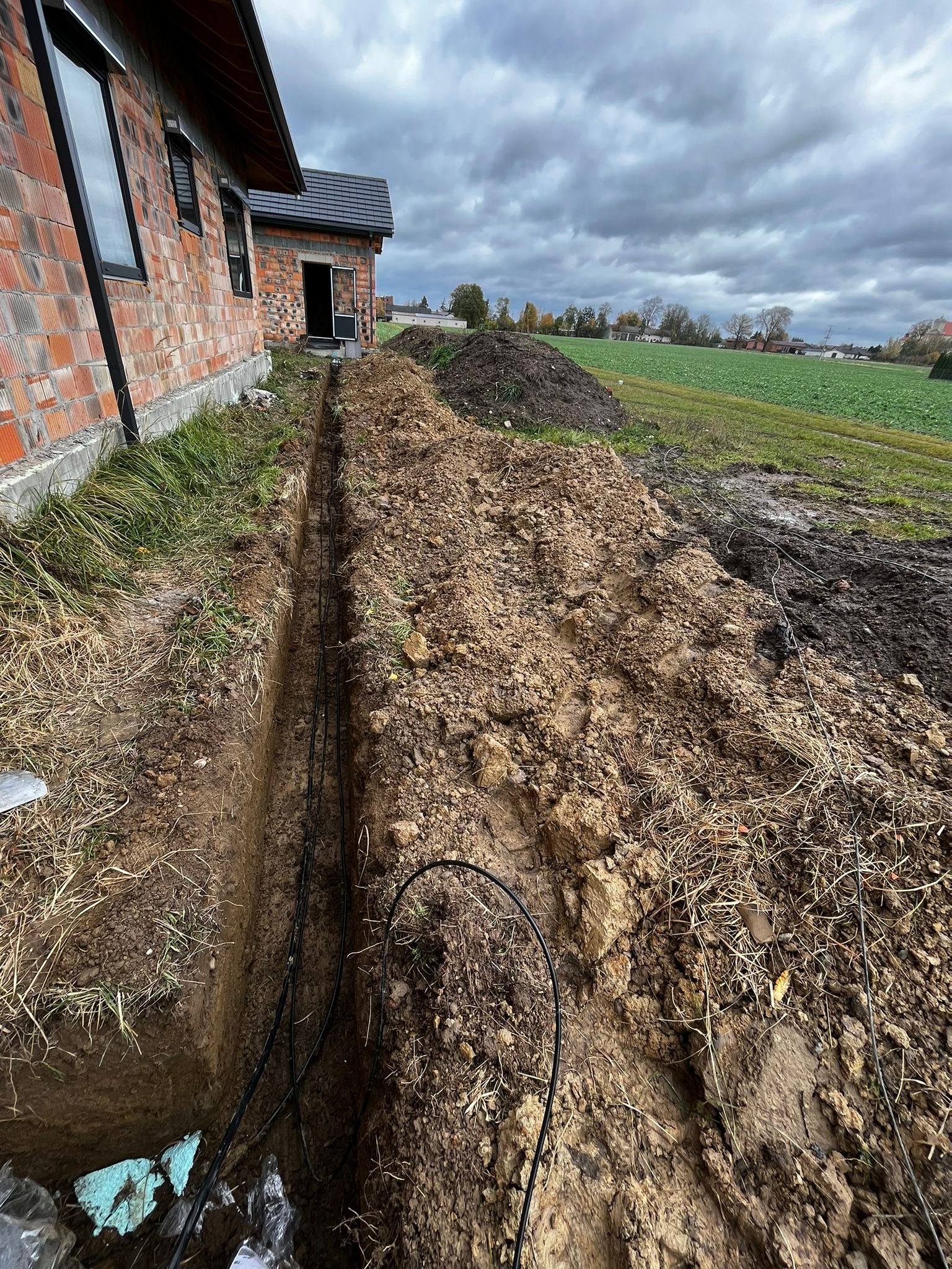 Wykop z ułożonymi kablami ziemnymi wzdłuż ściany budynku z czerwonej cegły, widok z góry na tle pochmurnego nieba i pola. Prace ziemne.