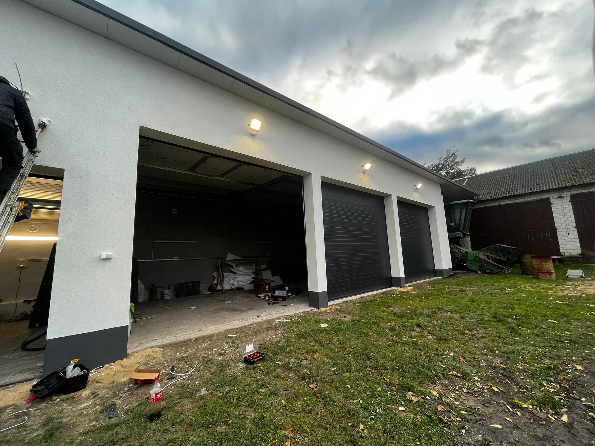 Nowoczesny garaż z trzema bramami segmentowymi w kolorze antracytowym. Na elewacji zamontowane lampy oświetleniowe. Osoba na drabinie wykonuje prace wykończeniowe.