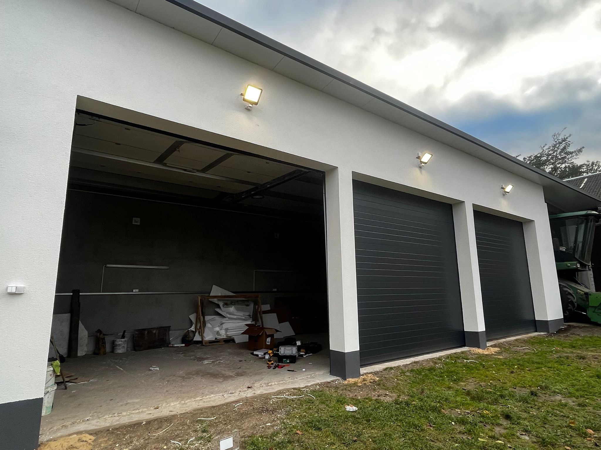 Nowoczesny garaż z trzema segmentowymi bramami w kolorze antracytu, oświetlony zewnętrznymi lampami. Jedna brama otwarta, widoczne narzędzia wewnątrz.