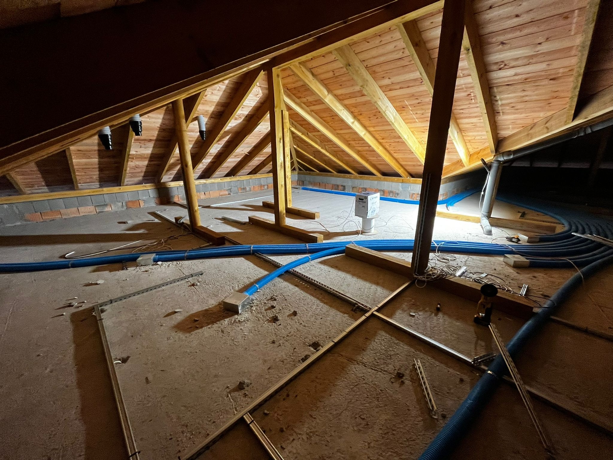 Poddasze w trakcie prac: widoczne drewniane elementy konstrukcyjne, niebieskie rury wentylacyjne, biała skrzynka i narzędzia. Ujęcie z dołu, ukazujące surowy stan poddasza.