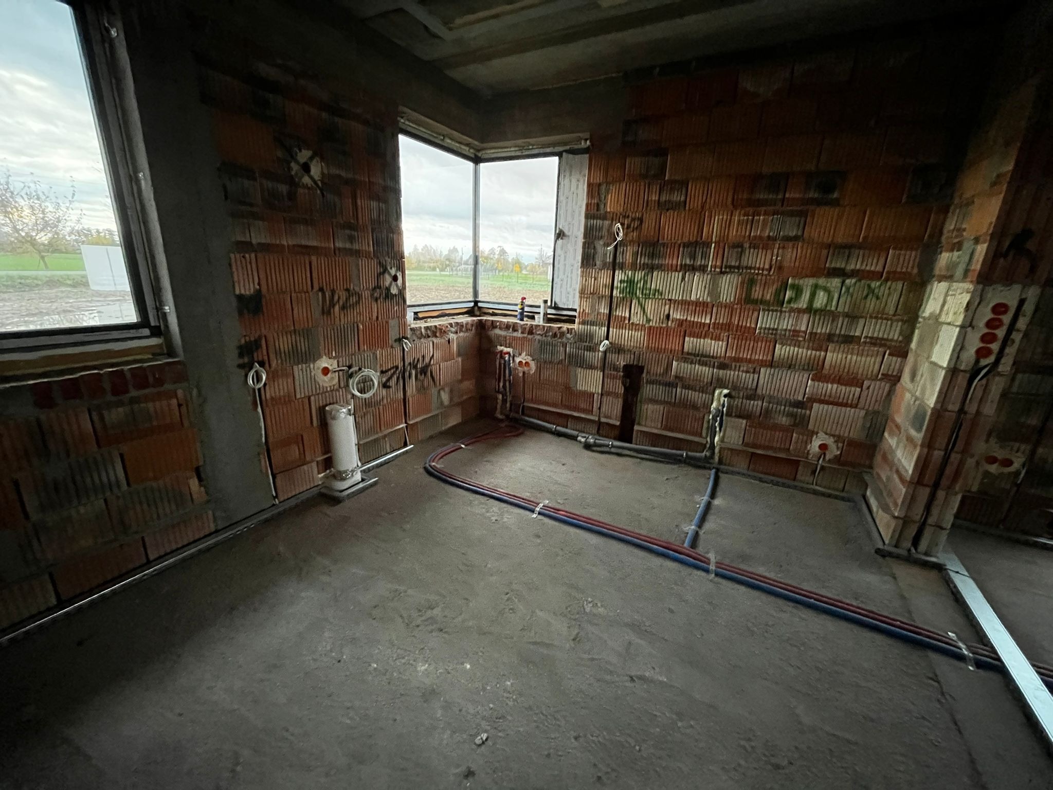 Surowy stan łazienki z widoczną instalacją wodno-kanalizacyjną w ścianach z cegły dziurawki i wylewką betonową. Okno z widokiem na zewnątrz. Etap prac instalacyjnych.